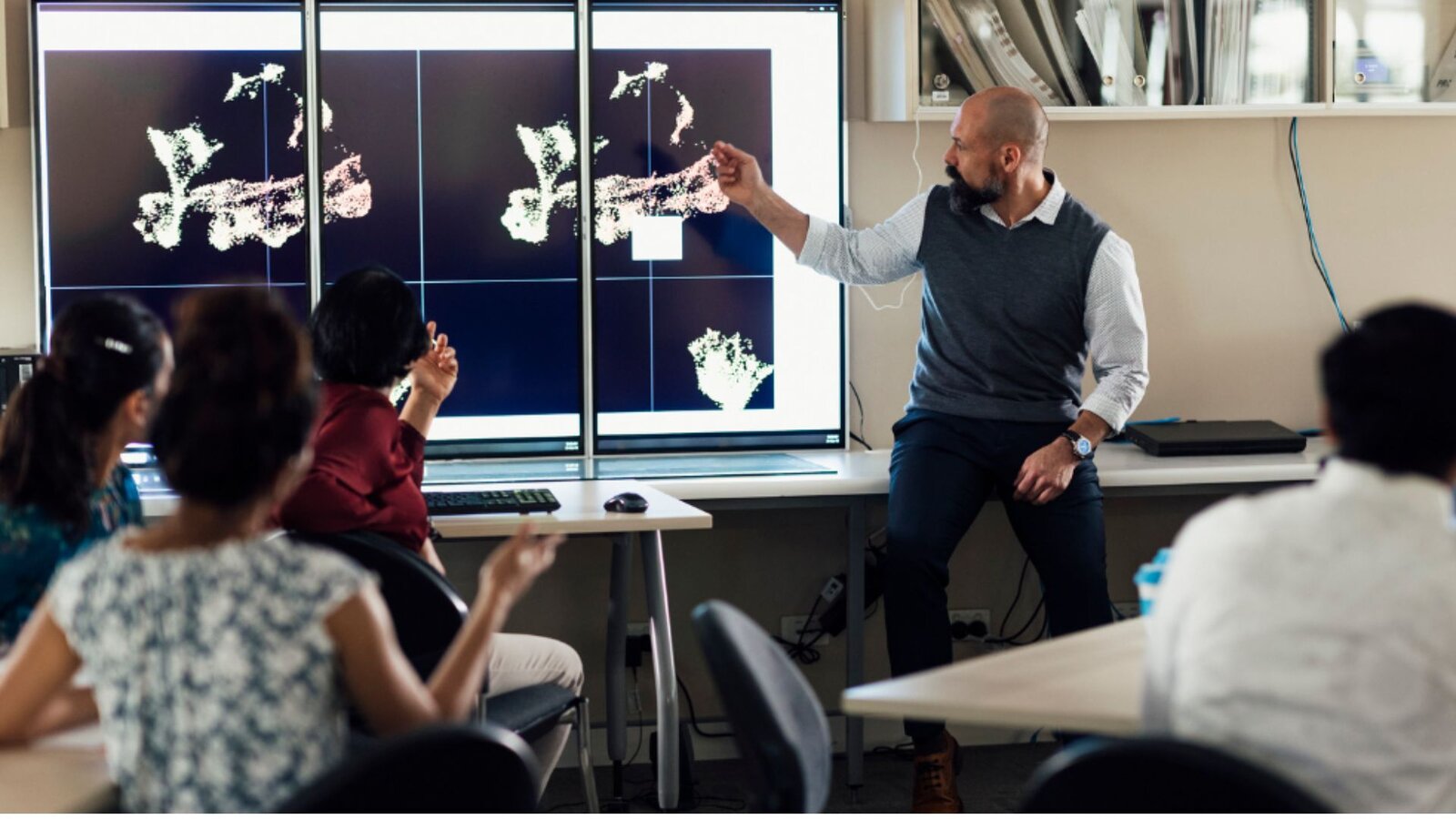 Researcher pointing at microscope images on large screens while colleagues around a table discuss them