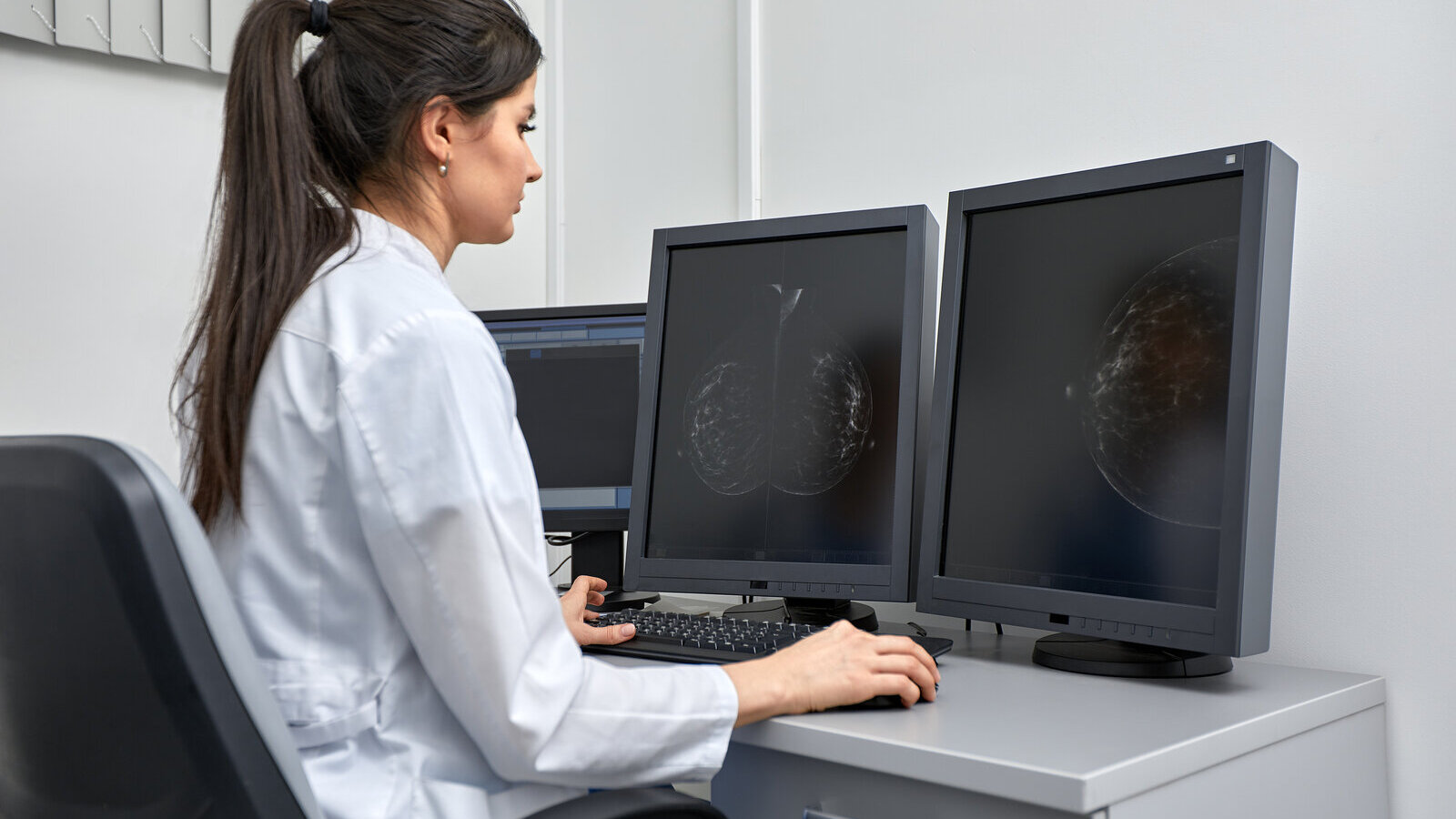 Clinician in a white coat reading images on two monitors at a workstation