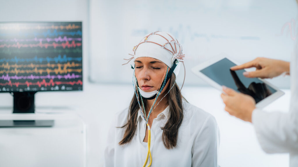 Adult wearing an EEG electrode cap while brain wave signals appear on a monitor