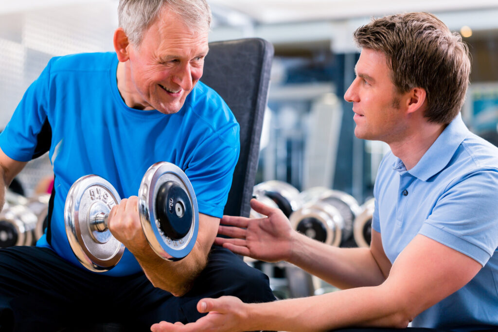 Older man lifting a dumbbell while a trainer guides him in a gym