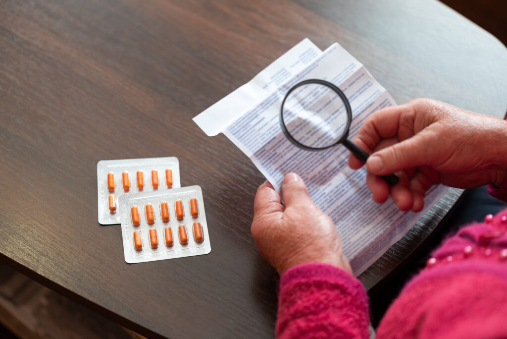 Hands holding a magnifying glass over a medicine leaflet next to blister packs of pills