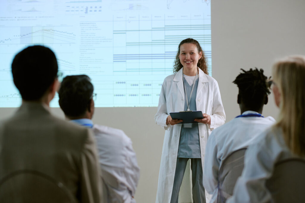A crowded slide behind the speaker Researcher in a lab coat speaking to colleagues in front of a projected slide crowded with charts and tables