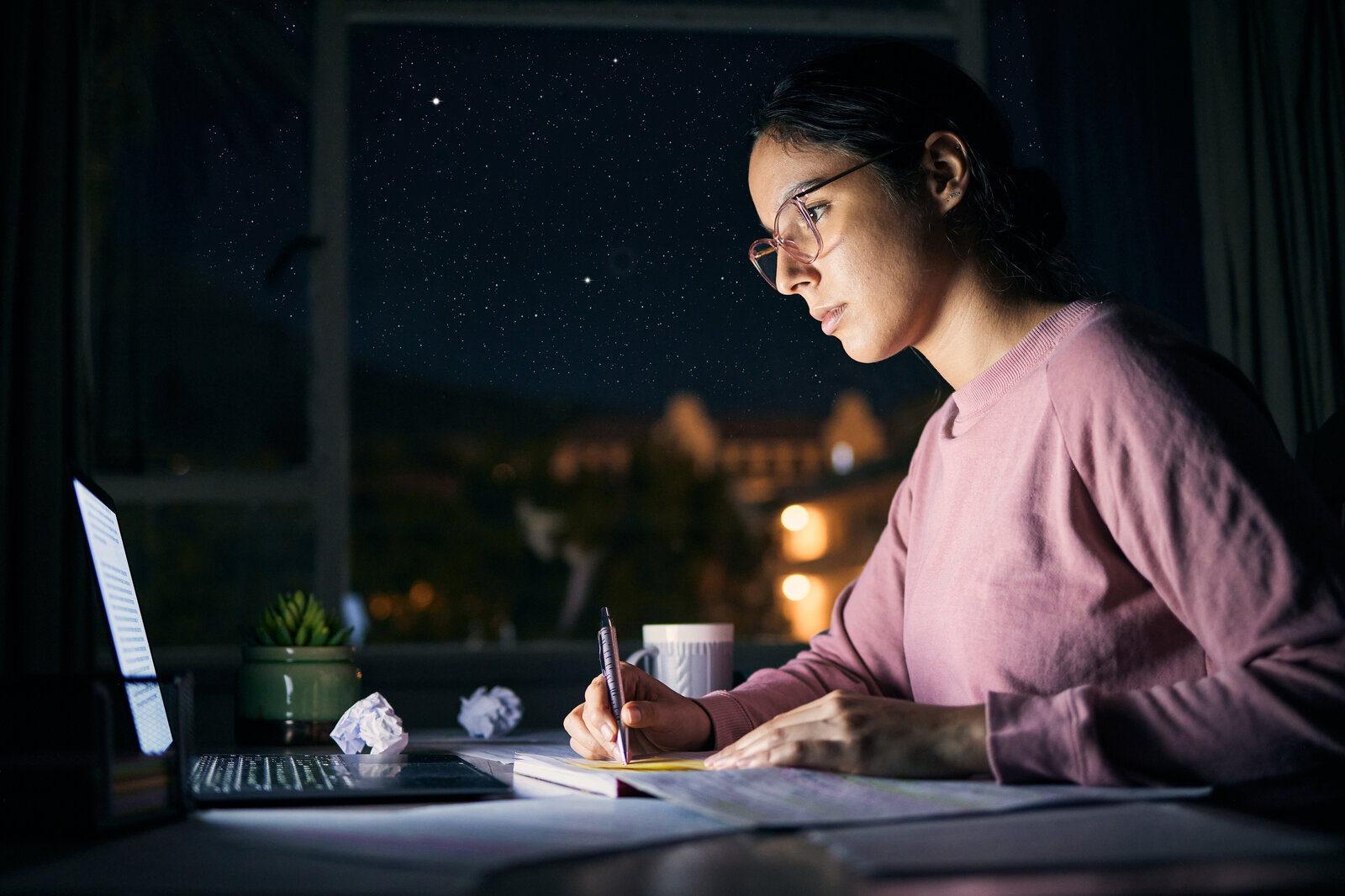 Researcher writing notes by lamplight next to a laptop at night