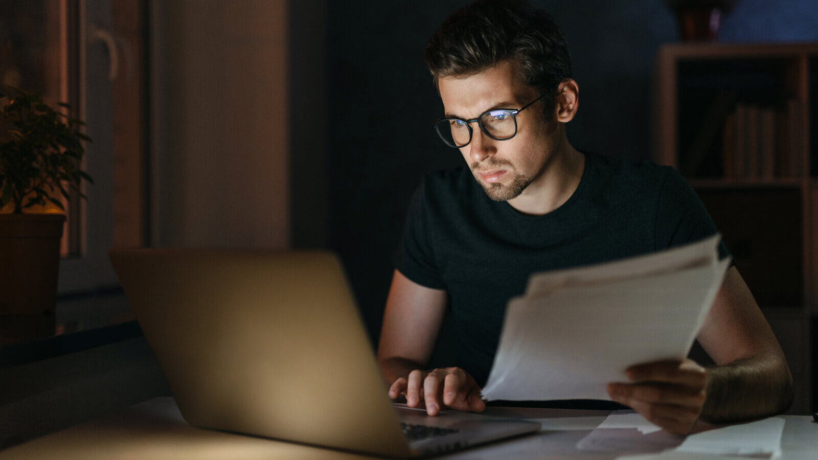 Person reading printed pages beside a laptop late at night