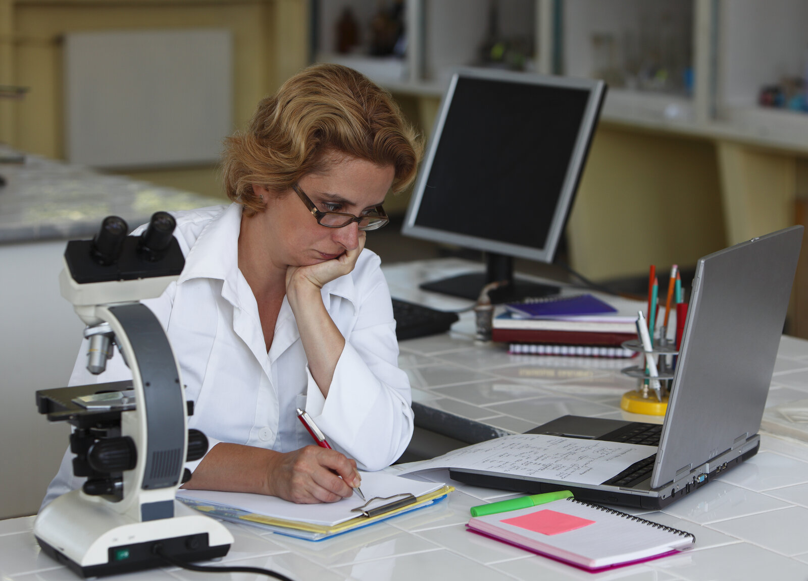 Scientist at a lab desk thinking over handwritten notes beside a laptop and microscope