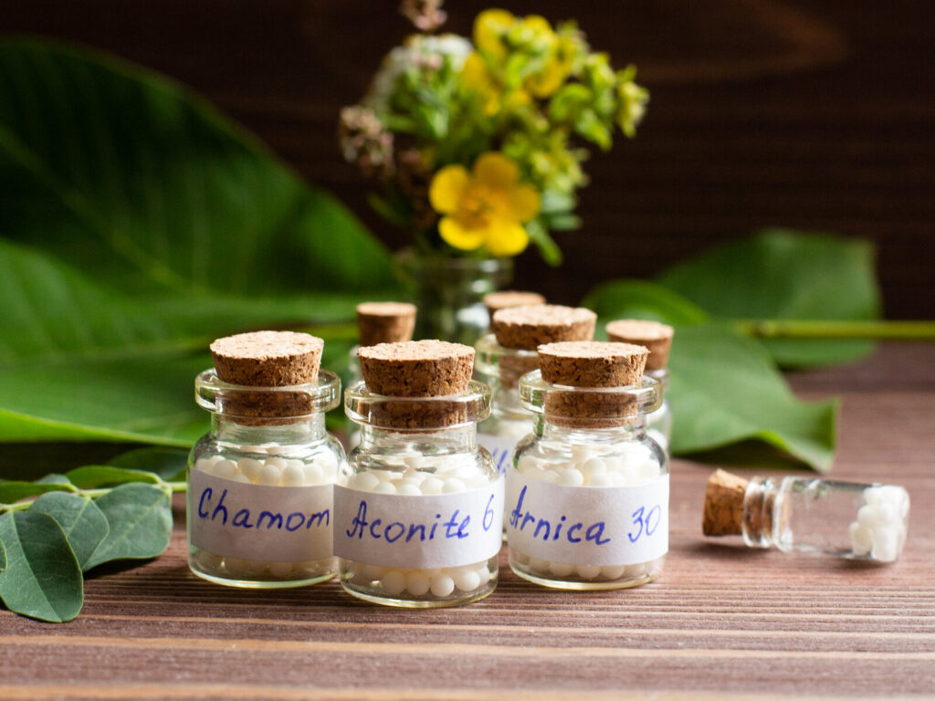 Small glass jars of white homeopathic pellets labeled Chamomilla, Aconite and Arnica on a wooden table