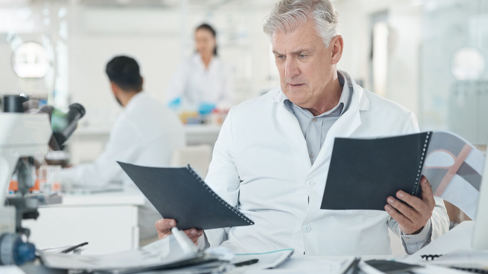 Older scientist in a white lab coat frowning as he reads two bound papers at a lab bench